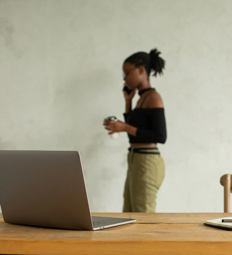 Kundenqualifizierung automatisieren - Solopreneurin steht vor einer Wand und telefoniert. Im Vordergrund ist ein Laptop auf einem Schreibtisch zu sehen.